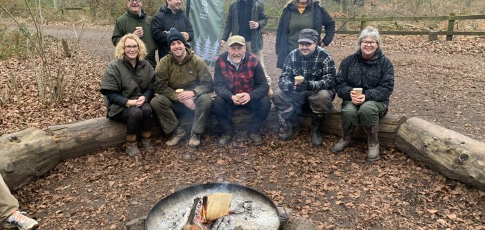 Group of people in a wooded area sat on a log in front of open fire