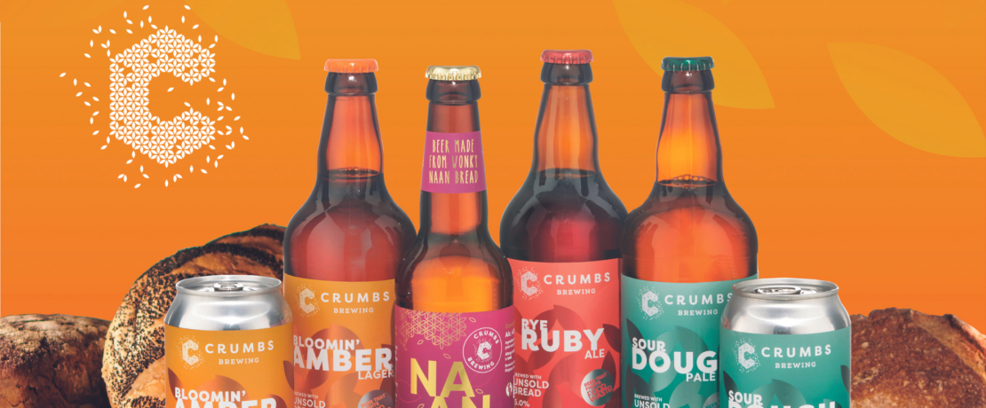 Image of 3 bottles and 2 cans of beer made by sustainable craft brewery, Crumbs Brewing. Bottles and can are on a flat surface against an orange background with loaves of bread behind them