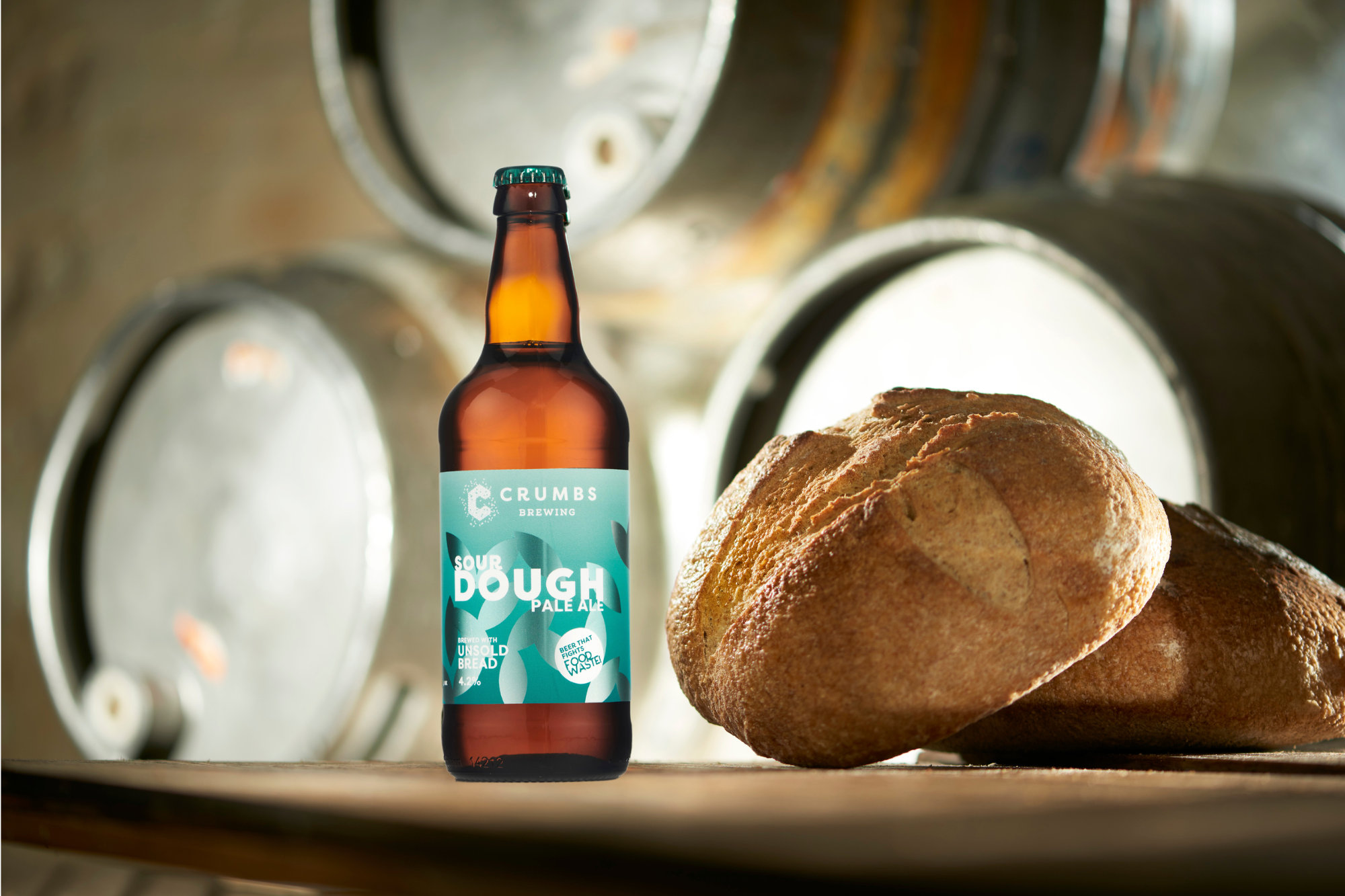 Image of a beer bottle made by sustainable craft brewery, Crumbs Brewing. The bottle is is on a flat surface with metal beer barrels stacked behind and a loaf of bread next to it.