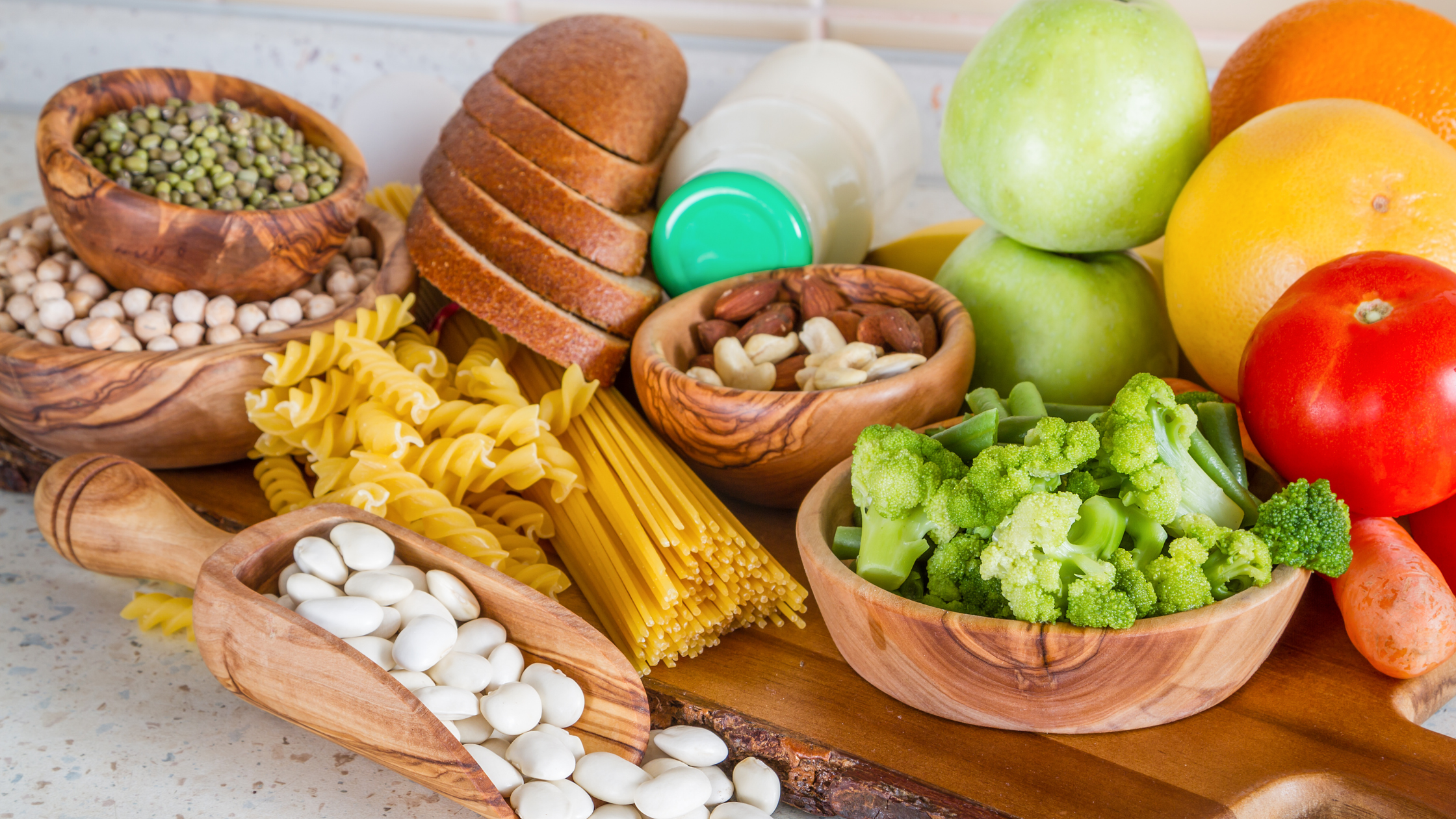 Image of selection of vegetarian foods on a wooden platter on a table. Foods include bread, pulses, pasta, fruit, veg and nuts